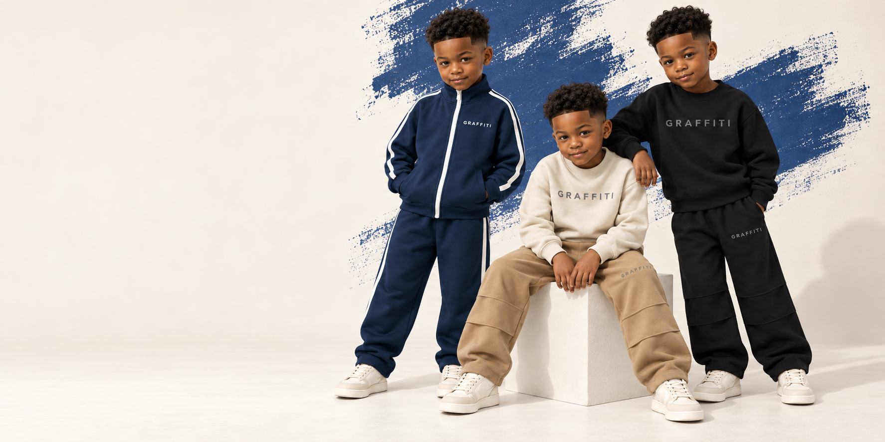 Three children modeling Graffiti Dreams sweat sets in navy, cream, and black against a cream wall with a royal blue paint stroke